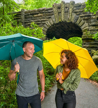 Two people walking in Central Park, holding The Original Duckhead umbrellas — one green, one yellow — and smiling under Glen Span Arch in Central Park.