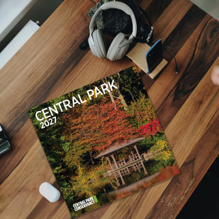 A 2027 wall calendar titled 'CENTRAL PARK' lies on a wooden desk, styled with a small potted plant, white over-ear headphones, a smartphone, and a white mouse. The calendar cover features a colorful autumn park scene with trees, a pond, and a pavilion.
