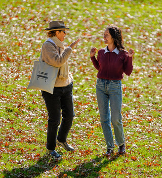 Two people standing in a grassy field in Central Park  NYC with fallen leaves, one holding a grey tote bag with a Central Park Bench printed on it.