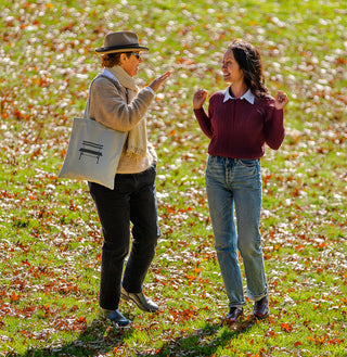 Two people standing in a grassy field with fallen leaves, one holding a tote bag featuring a Central Park bench. 