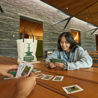 Two people playing cards at a table with a Central Park Conservancy tote bag in the background.