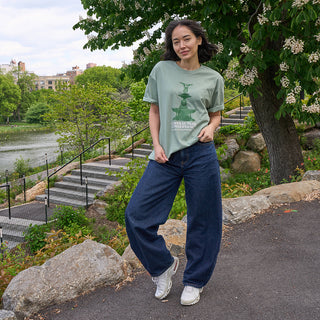 Person wearing a green t-shirt with a print of Bethesda Fountain in Central Park and the Central Park Conservancy wordmark and blue jeans standing near the Davis Center among trees and the Harlem Meer in the background.