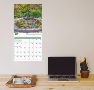 A vertical wall calendar page featuring a July garden pond photograph above a printed calendar grid, displayed on a beige wall above a wooden desk with a laptop, cactus, and tray.